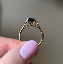 Gold ring with a green gemstone held by a hand with pink nail polish against a blurred background